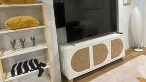 Una sala de estar con un televisor de pantalla plana grande montado en una pared de ladrillo blanco, estantes de madera con almohadones decorativos y un altavoz blanco.