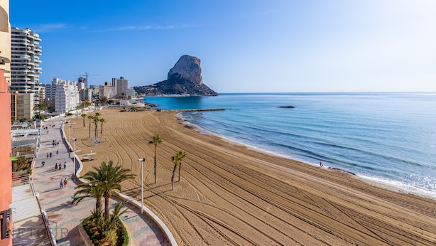 A beach with a prominent rock formation, a promenade with palm trees, and buildings in the background. - Inftour Oceanic 6 Apartamento (Calp)