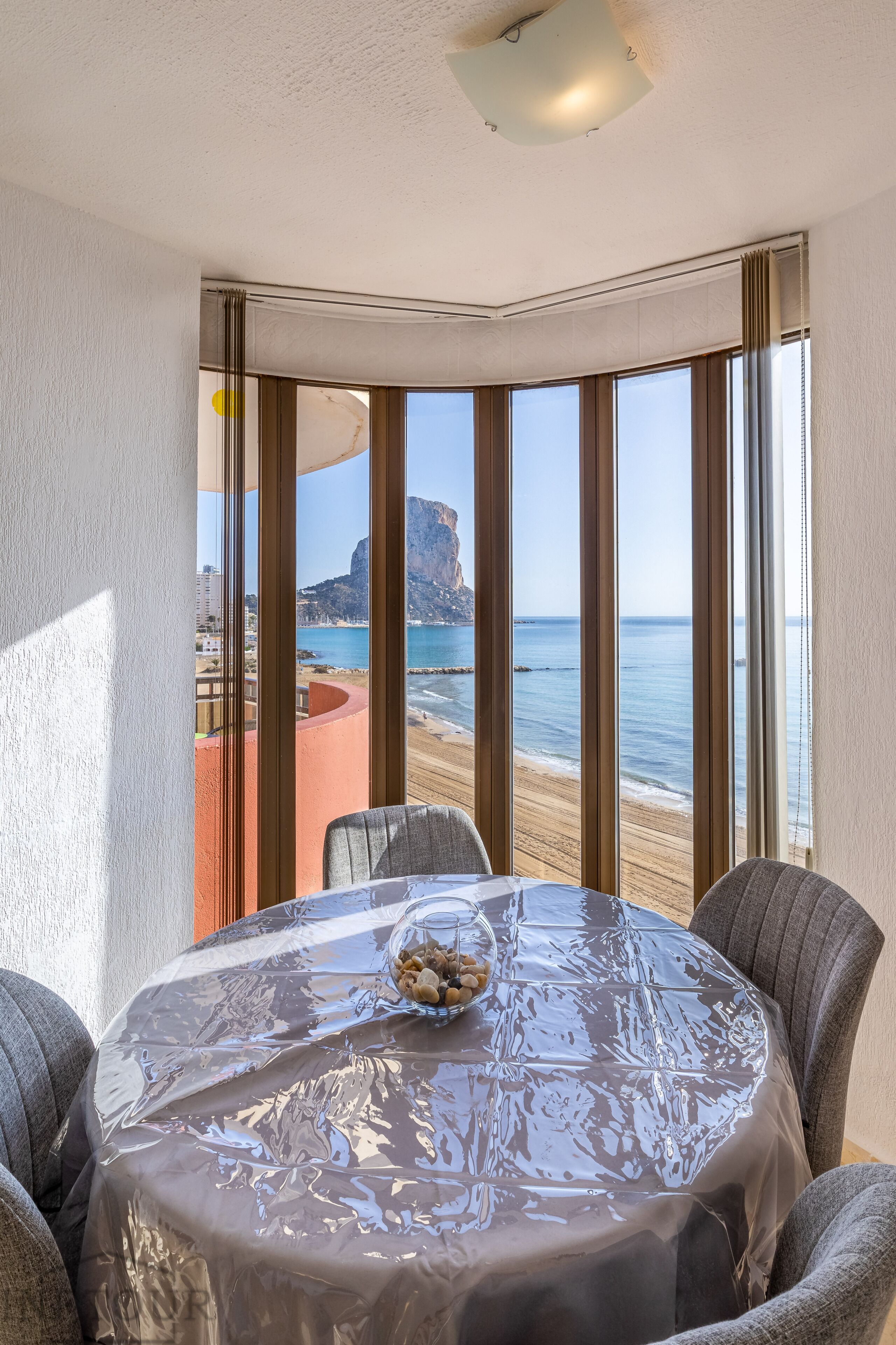 A dining area with a table covered in a clear plastic tablecloth, two chairs, and a bowl of nuts. Large windows offer a view of the beach and ocean.