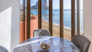 A dining area with a table covered in a clear plastic tablecloth, two chairs, and a bowl of nuts. Large windows offer a view of the beach and ocean. - Inftour Oceanic 6 Apartamento (Calp)