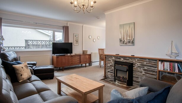 A living room with a sofa, ottoman, coffee table, fireplace, television, bookshelf, and a view of the outside. - Glan Heulog - 3 Bed Holiday Home - Saundersfoot (Saundersfoot)