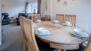 A dining area with a wooden table set for four, surrounded by wooden chairs. There is a window with curtains, a radiator, and wall-mounted plates. - Glan Heulog - 3 Bed Holiday Home - Saundersfoot (Saundersfoot)