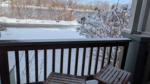 Eine Holzdielen-Terrasse mit zwei metallenen Tischen, die mit Schnee bedeckt sind, mit Blick auf eine verschneite Landschaft, kahle Bäume und ein weit entferntes Haus.
