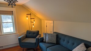 Ein Wohnzimmer mit blauem Sessel, blauem Sofa, gemusterten Kissen, einer Stehlampe mit freiliegenden Glühbirnen und einer Heizung.
