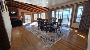A spacious dining area with a wooden table and chairs, a large rug, and a view of the outdoors through large windows. - Sunlit Modern Retreat with Wraparound Deck & Sustainable Design (Oakhurst)
