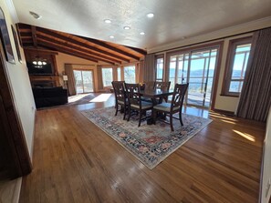 A spacious dining area with a wooden table and chairs, a large rug, and a view of the outdoors through large windows. - Sunlit Modern Retreat with Wraparound Deck & Sustainable Design (Oakhurst)