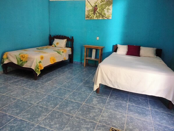 Una habitación con dos camas, una con cubrecama floral y la otra con cubrecama blanca lisa. Hay una mesita de noche de madera con una lámpara y un cuadro enmarcado en la pared.