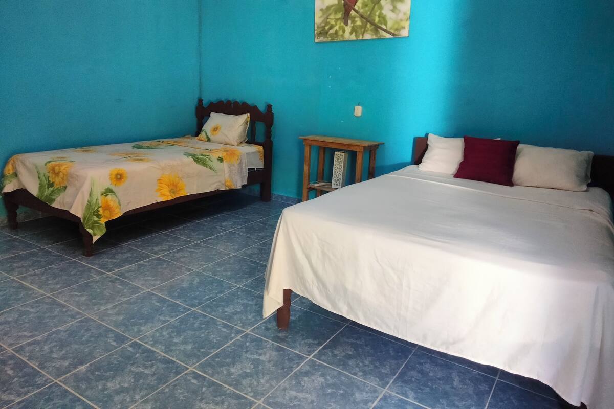 A room with two beds, one with a floral bedspread and the other with a plain white bedspread. There is a small wooden table and a wall-mounted shelf.