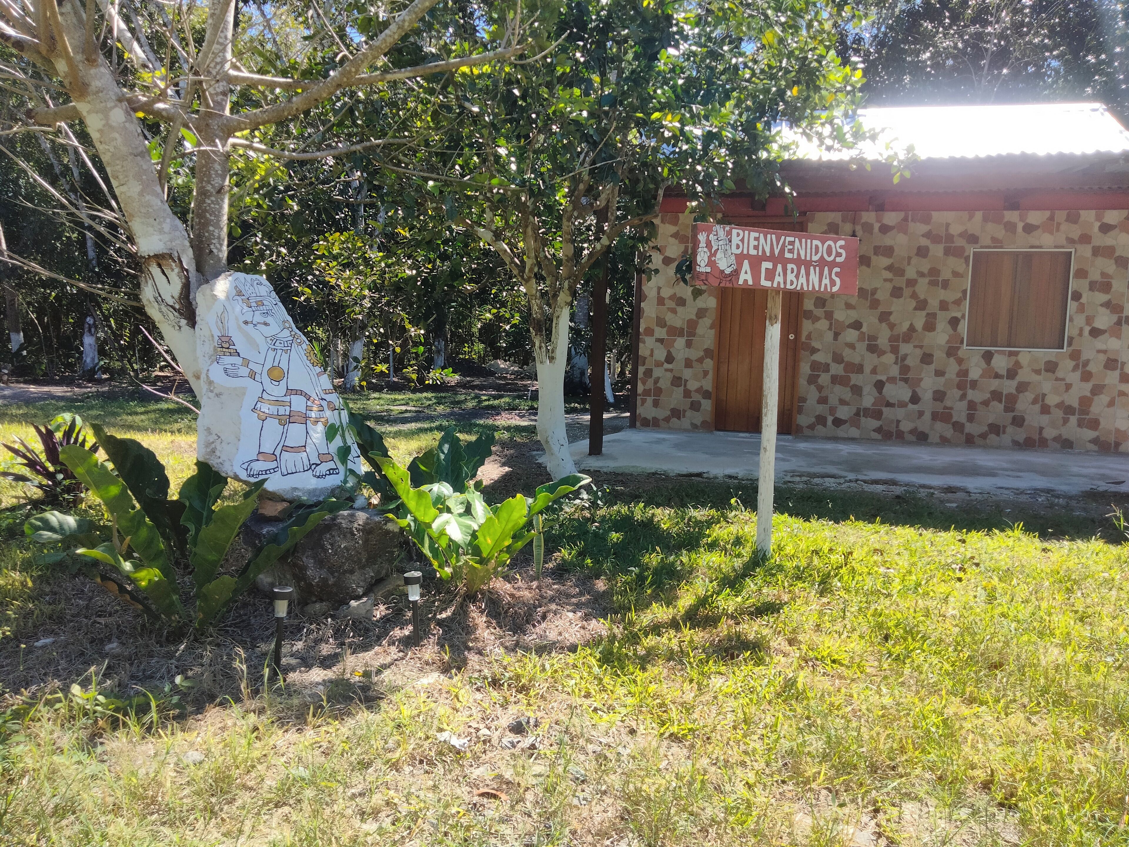 A sign welcoming visitors to a location, a stone building with a tiled facade, and a tree with a decorative sign.