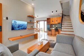 Un salon moderne doté d’un téléviseur à écran plat, de canapés gris, d’étagères en bois et d’un escalier menant à un espace cuisine.