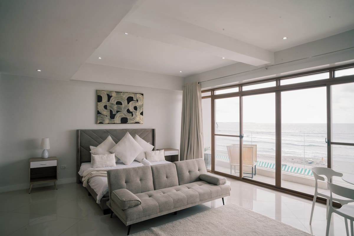A modern bedroom with a large bed, a sofa, a nightstand, and a view of the ocean.
