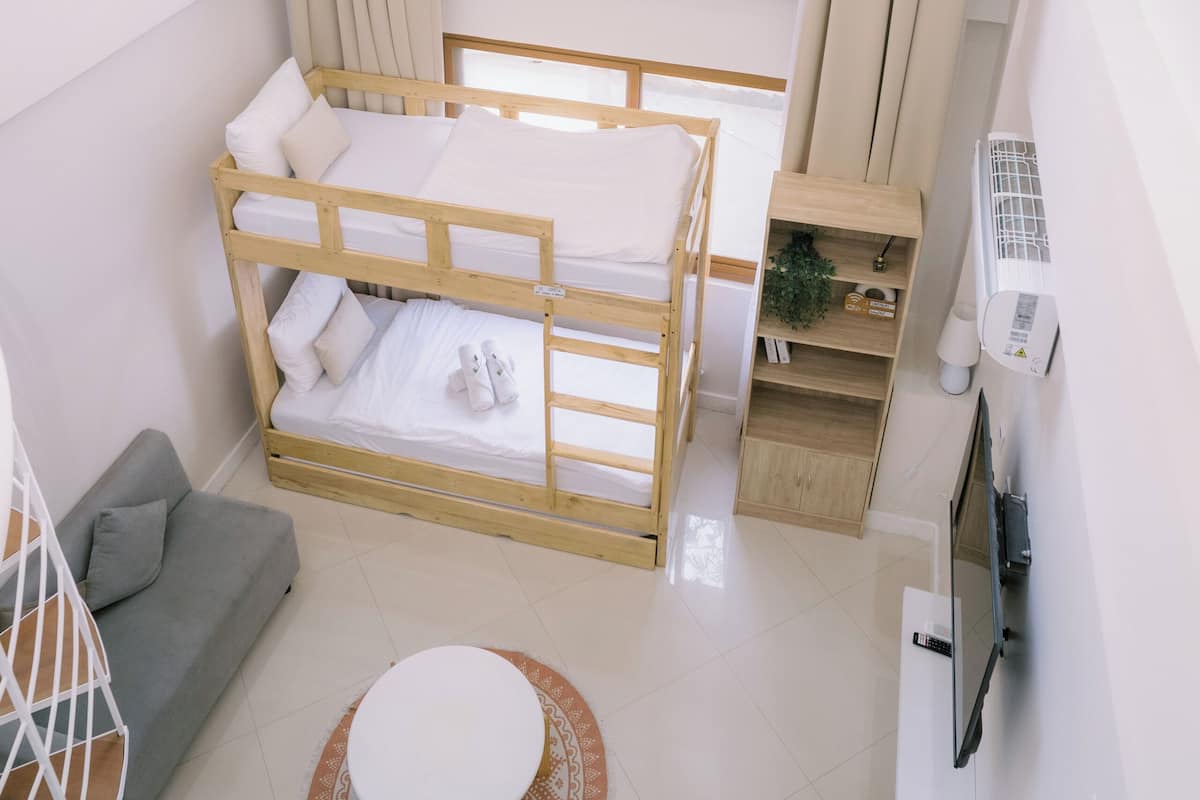 A bedroom with a bunk bed, a grey sofa, a round white table, a TV, and a window with curtains.