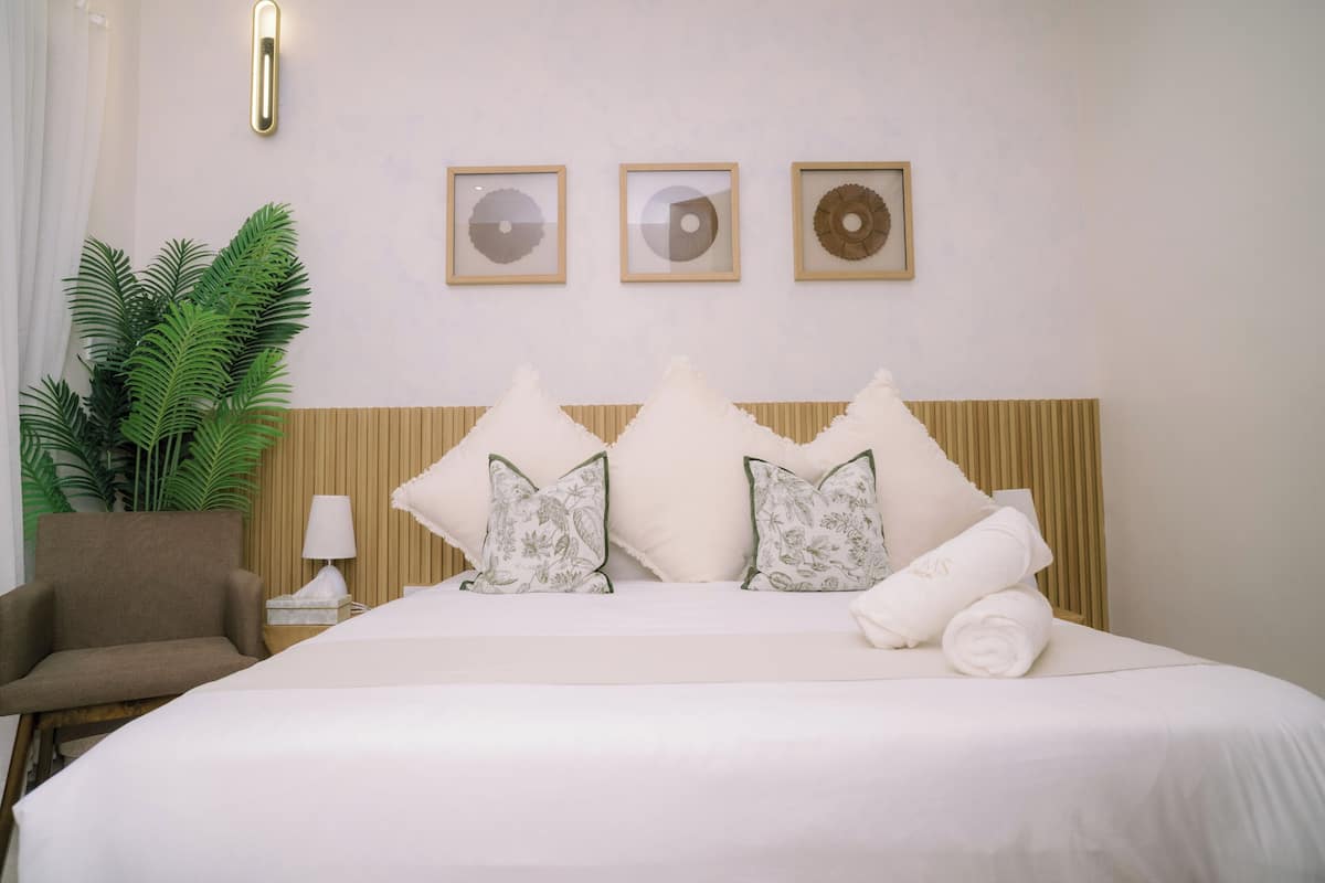 A hotel room with a bed, pillows, a chair, a lamp, a plant, and three framed wall decorations.