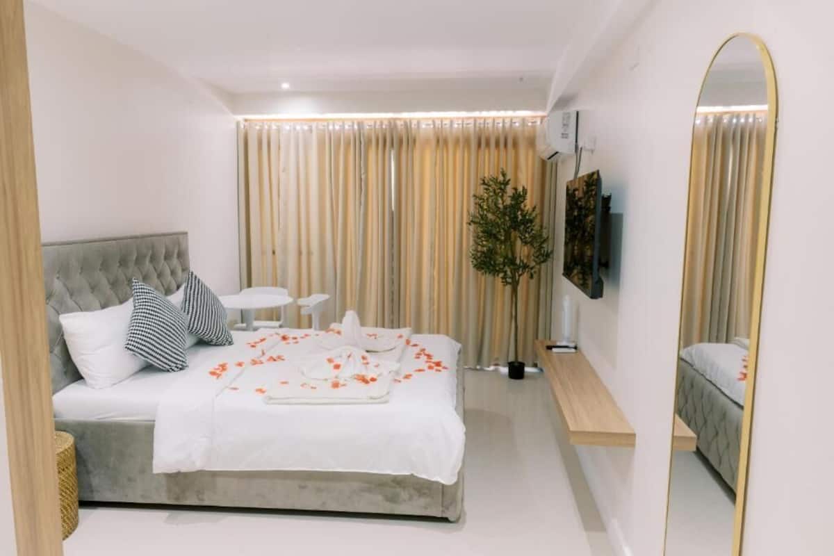 A modern hotel room with a large bed, a bedside table, a small desk, a mirror, and a potted plant.