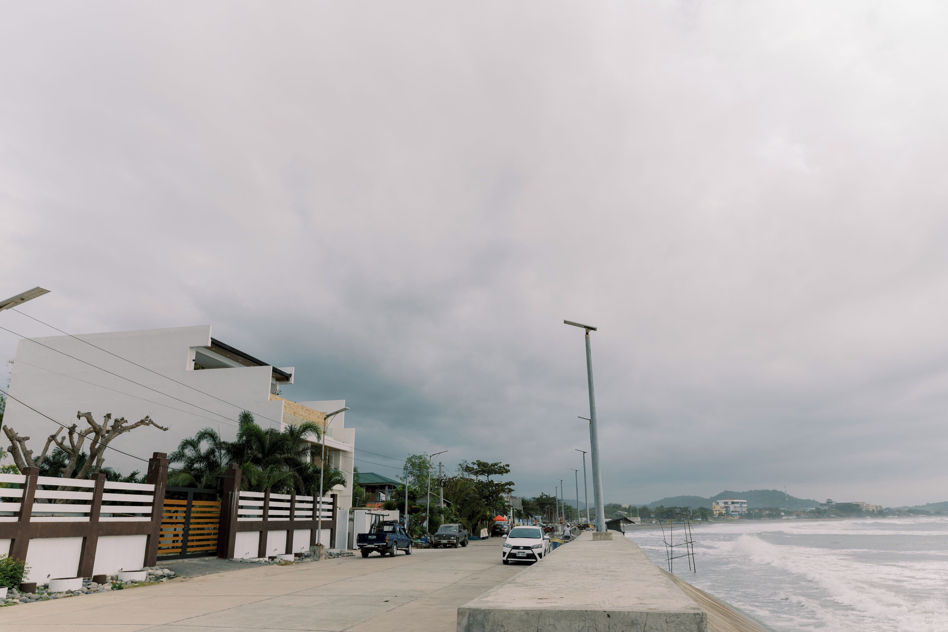 Una calle costera con edificios modernos, coches aparcados y una playa al fondo.