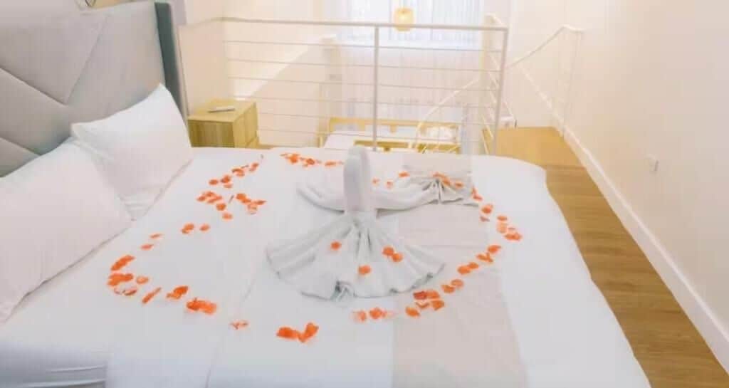 A neatly made bed with white linens and orange flower petals arranged in a heart shape.