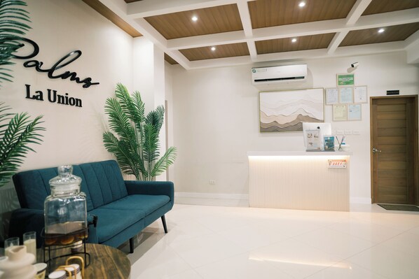 A reception area with a teal sofa, a glass jar on a wooden table, and a reception desk. - Palms La Union Suites (San Juan)