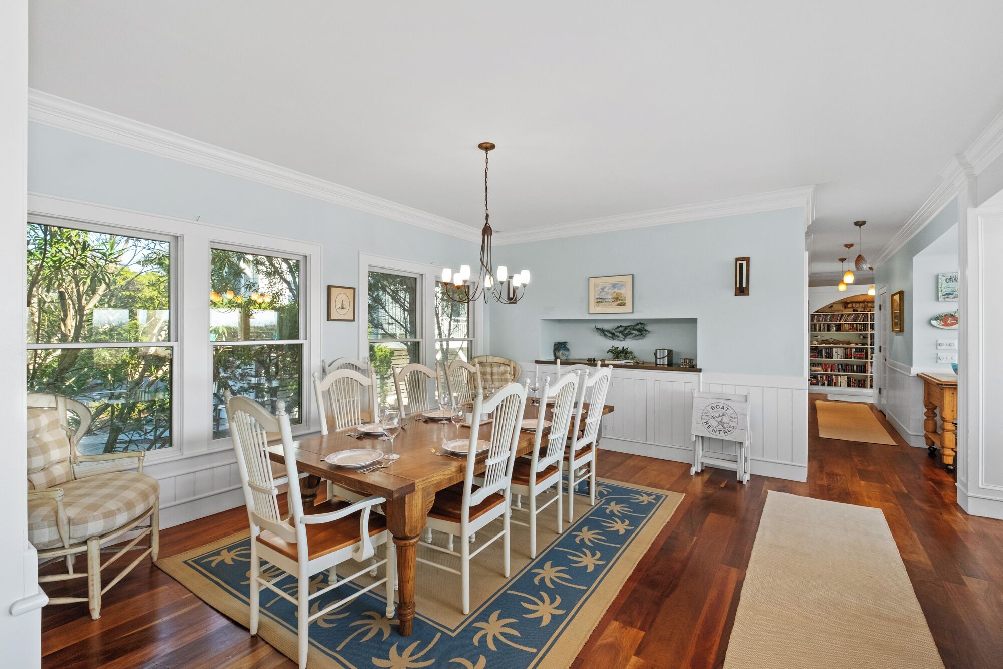 A dining area with a wooden table and white chairs, large windows with a view of greenery, a kitchen with white cabinets, and a bookshelf filled with books.