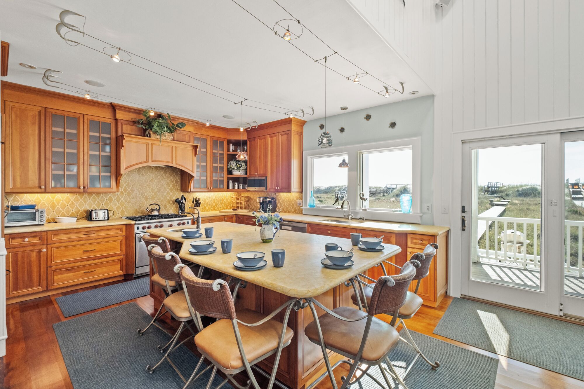 A spacious kitchen with wooden cabinets, a central island with chairs, a stove, and a view through large windows.