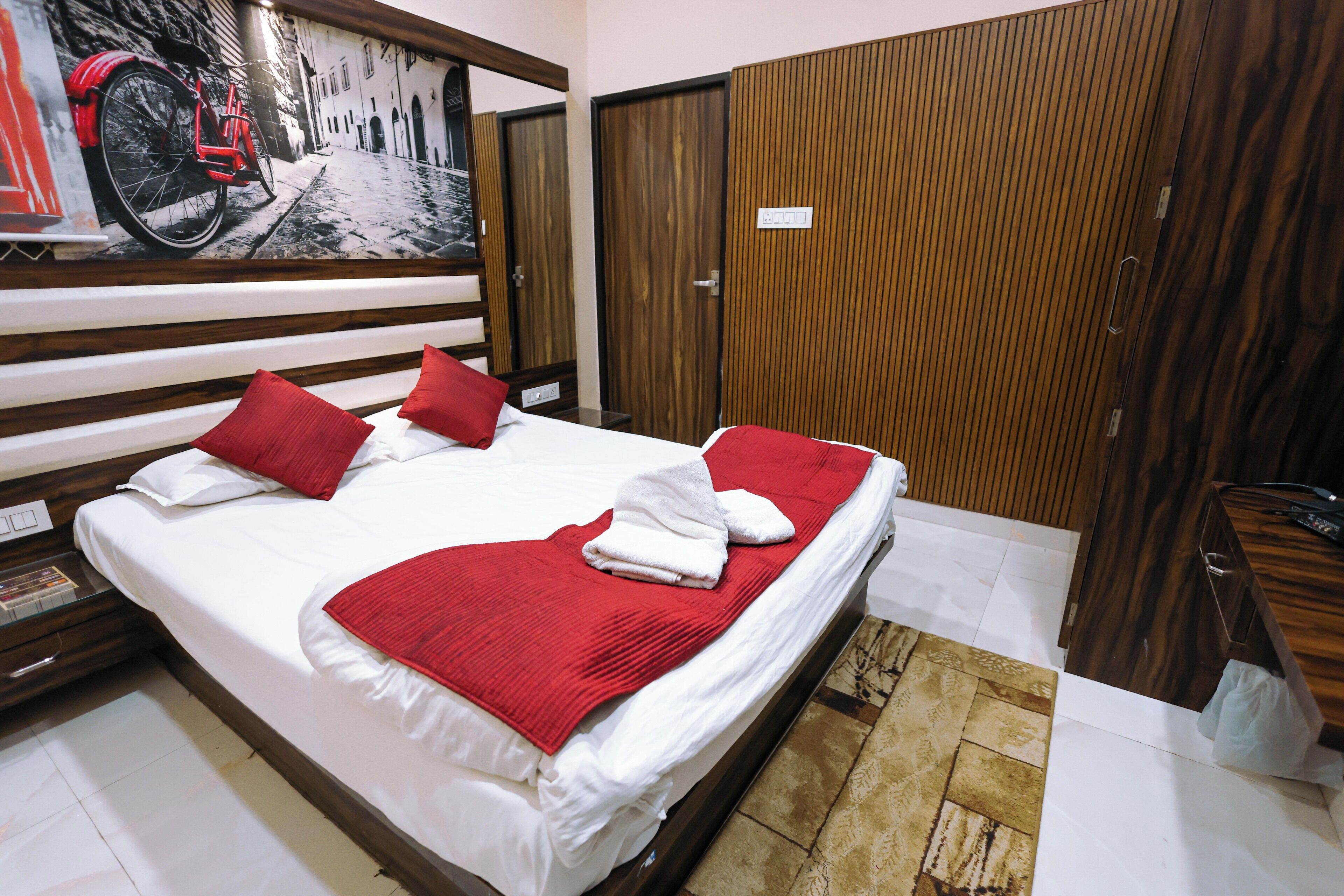 A hotel room with a large bed, wooden headboard, red pillows, a red blanket, and a painting of a bicycle on the wall.