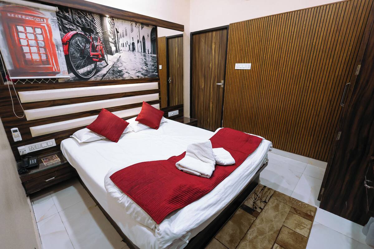 Une chambre d’hôtel comprenant un lit, un ensemble d’oreillers rouge et blanc, une couverture rouge, une tête de lit en bois, un téléphone fixé au mur et une porte ornée d’un panneau en bois.