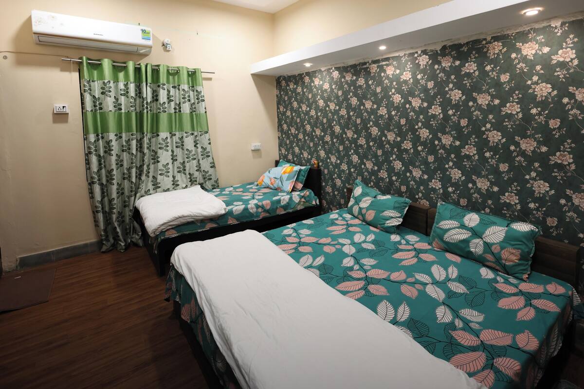 Une chambre avec deux lits, une couvre-lit à motifs floraux verts et blancs, un rideau vert et un papier peint à motifs floraux.