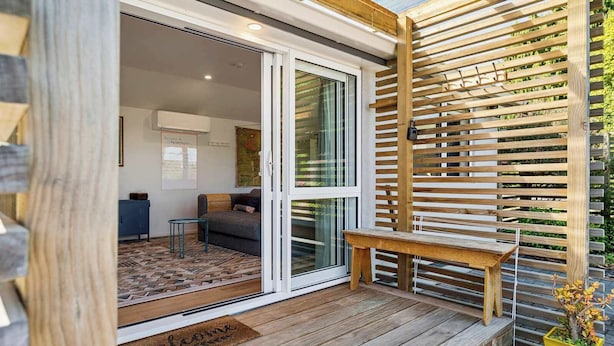 Una terraza de madera con un banco, un ventanal corredero que da a una sala de estar con un sofá y una mesa de centro, y una pequeña planta en maceta en la terraza.