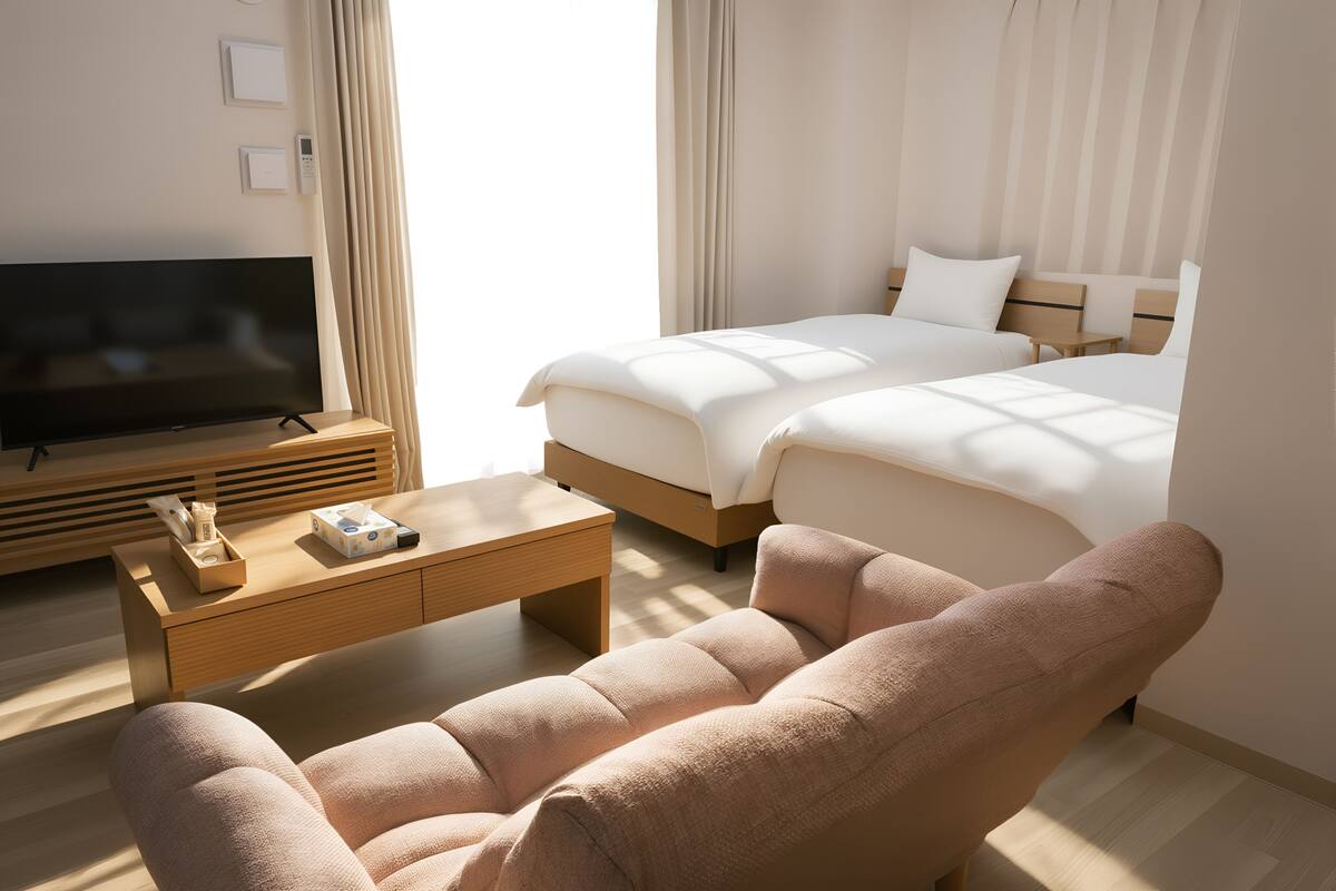 A hotel room with a flat-screen TV, a sofa, a bed with two pillows, a bedside table, and a window with curtains.