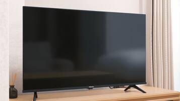 Un téléviseur à écran plat, posé sur un meuble en bois, avec une télécommande, est placé contre un mur orné de rideaux.