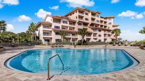 Una piscina con una escalera metálica, rodeada por un solárium con baldosas, y un edificio de varios pisos con balcones al fondo.