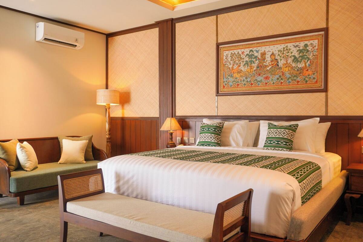 Quarto de hotel com uma cama grande, um sofá, uma poltrona, duas mesas de cabeceira com abajures e um ar condicionado fixado na parede.