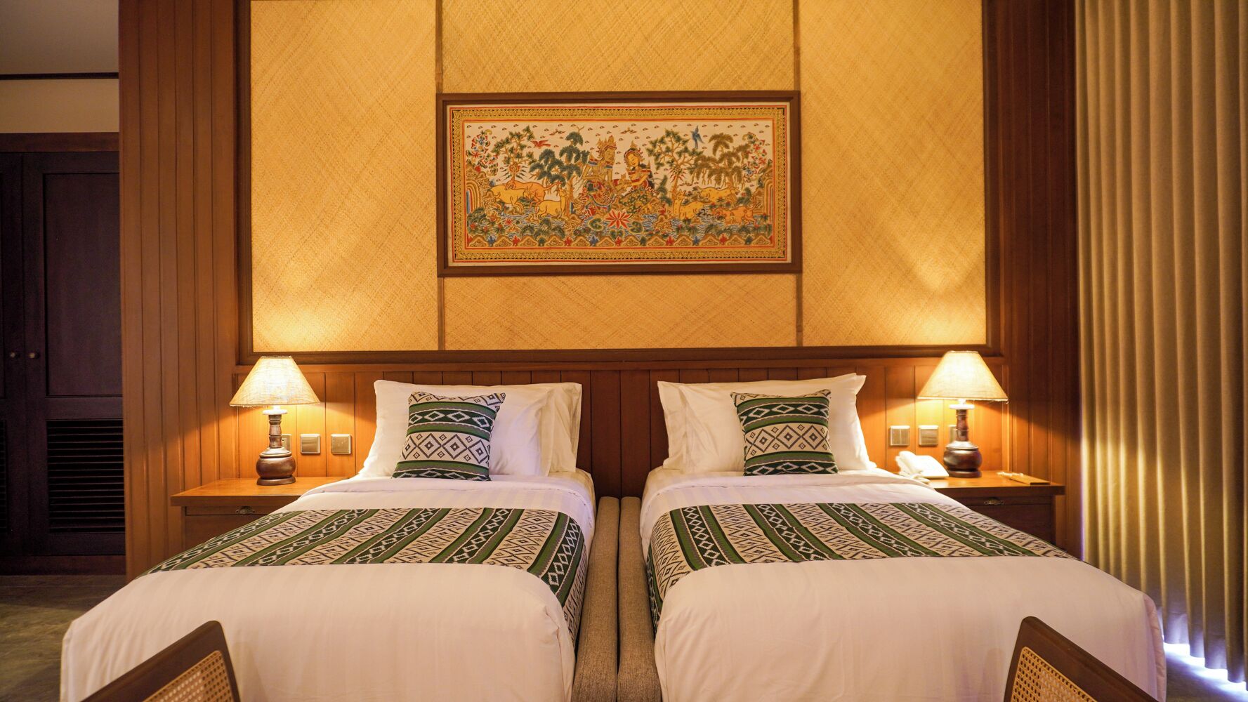 Quarto de hotel com duas camas, cada uma com colcha e almofadas estampadas, ladeadas por mesas de cabeceira com abajures. Uma obra de arte emoldurada está pendurada acima das camas.