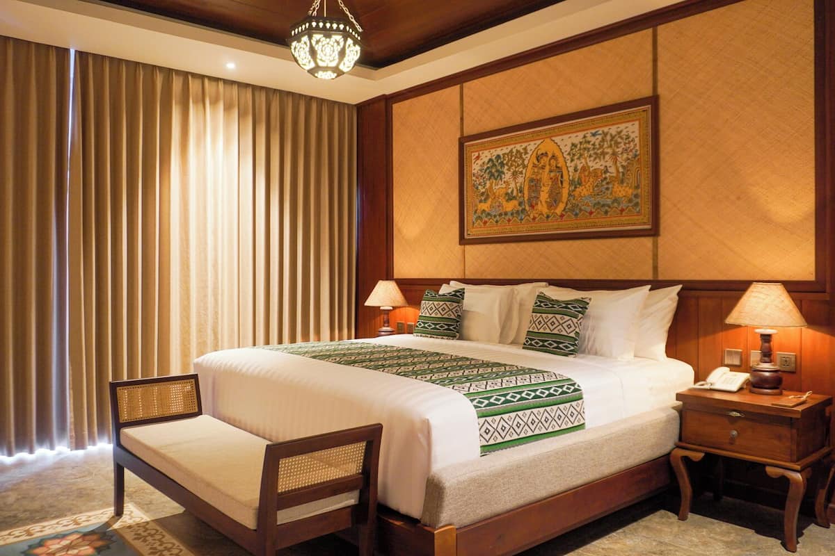 Quarto de hotel com uma cama grande, duas mesas de cabeceira, uma cadeira, um painel decorativo e uma cortina.