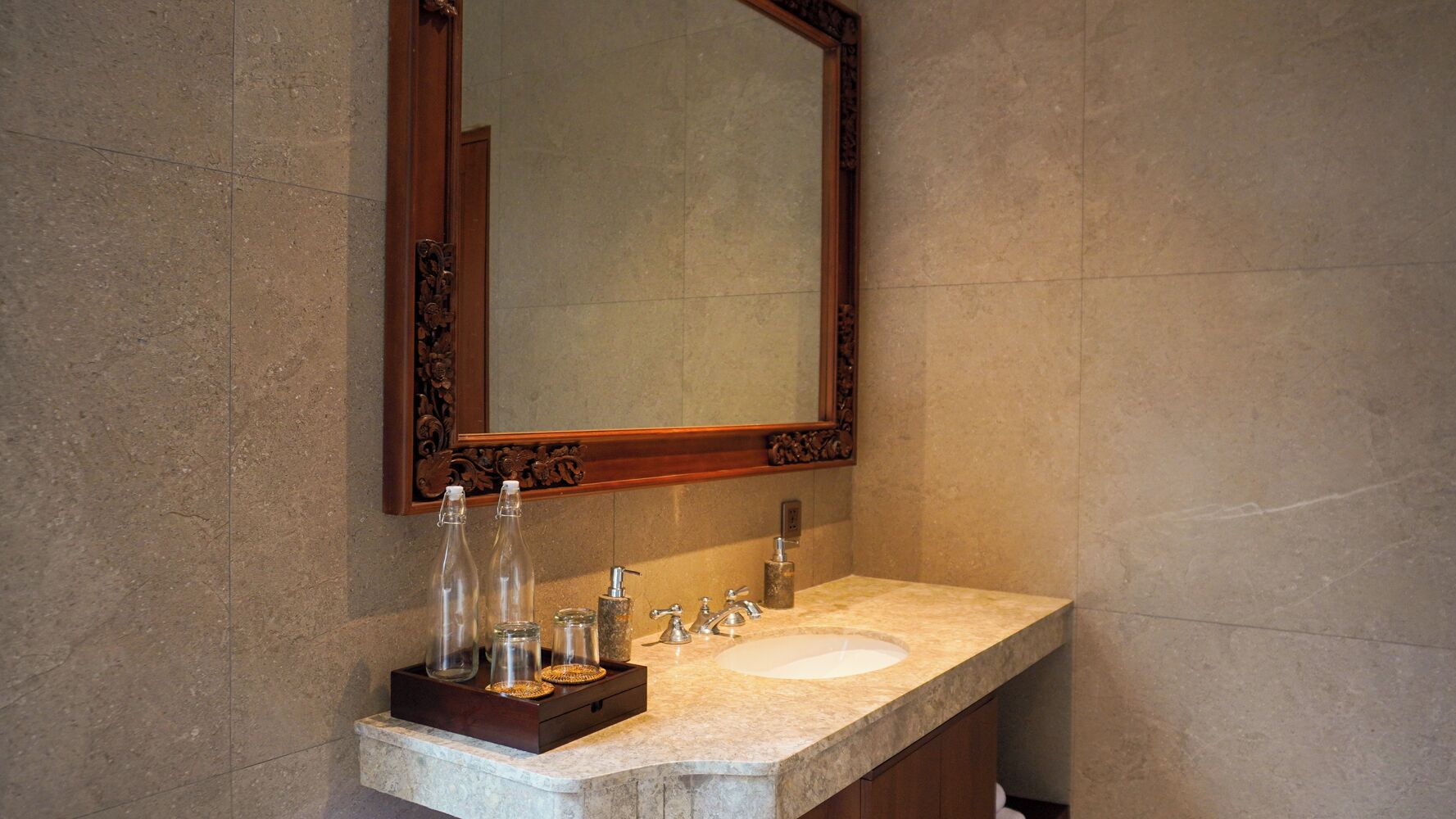 Une salle de bain avec un lavabo en marbre, un plateau en bois contenant des flacons et un distributeur de savon, et un grand miroir dot é d’un cadre orné.