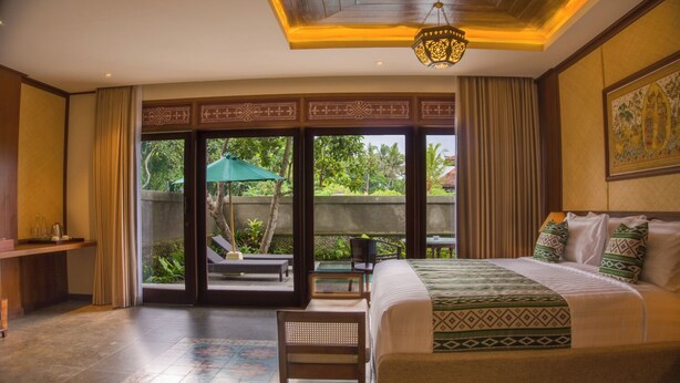 A hotel room with a large bed, a desk, and a view of a garden through sliding doors. - Naata Villas Ubud (Sukawati)