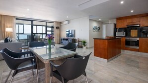 Una cocina moderna con una isla central, un comedor con mesa de vidrio y una sala de estar con sofá y televisor.