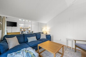 Ein kompakter Wohnraum mit Sofa, einem Holztisch, einem an der Wand montierten Fernseher und einer Küchenzeile im Hintergrund.