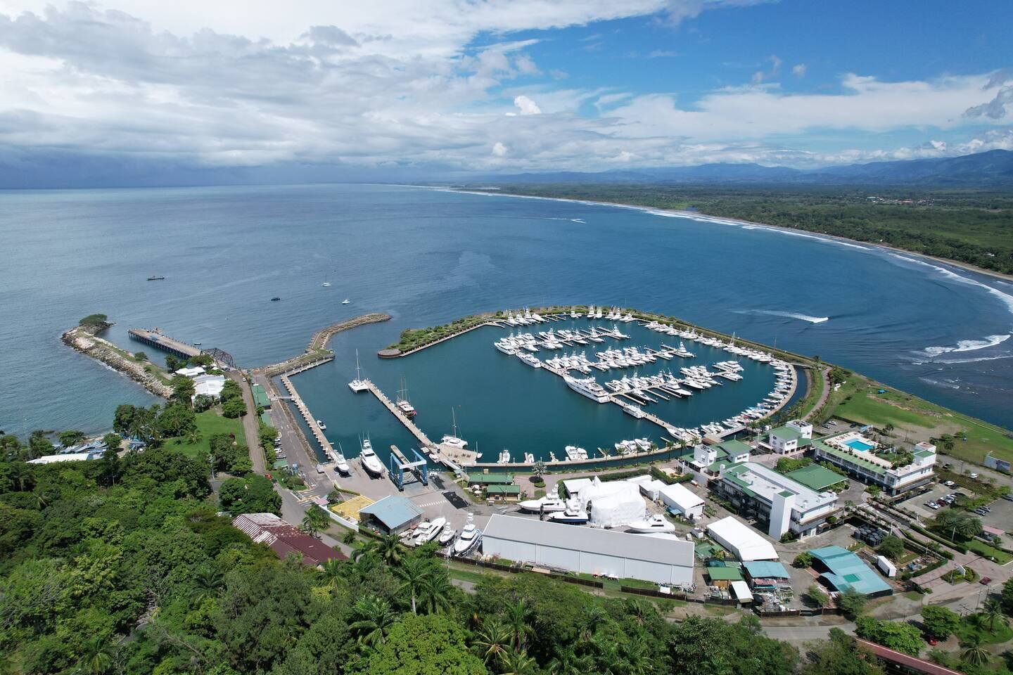 An aerial view of a marina with boats docked, surrounded by lush greenery and a clear sky.