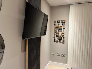 Ein modernes Wohnzimmer mit flachbildfernseher an der Wand, einem geometrischen Kunstwerk aus Holz und einem Fenster mit Jalousien.