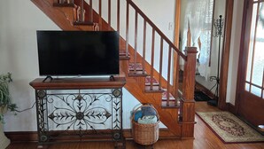 Een woonkamer met een houten trap, een televisie op een dressoir, een getextureerd vloerkleed en een rieten mand.