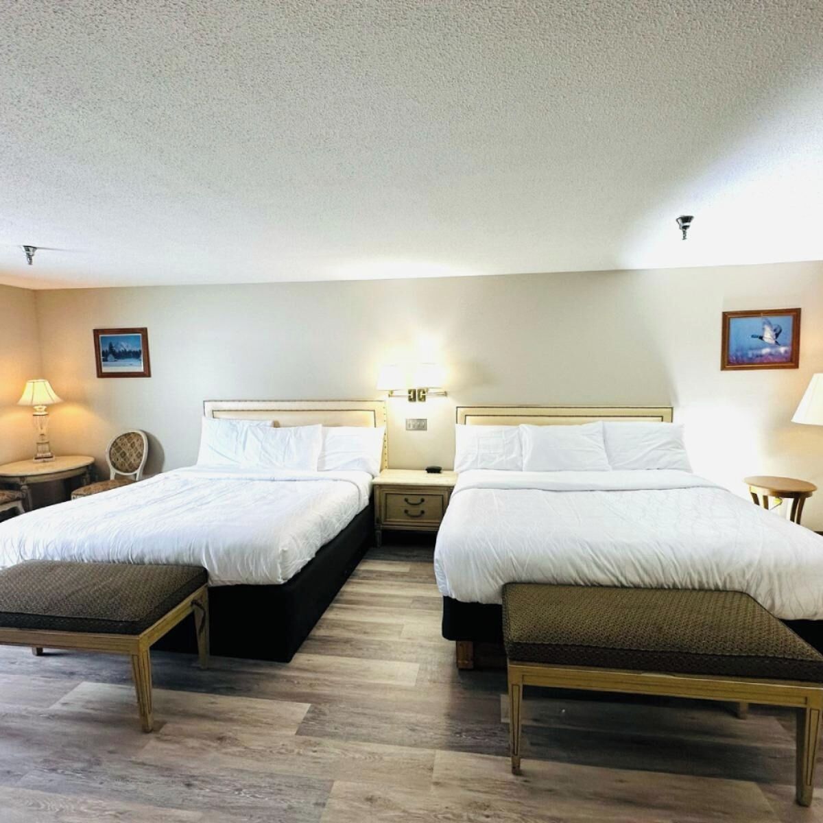 A hotel room with two beds, a small table with a lamp, a chair, and a bench.