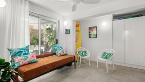 A modern room with a bed, two white chairs, a surfboard, and a tropical-themed painting. - Seaspray Unit 2, 21 Warne Tce, Kings Beach (Kings Beach)