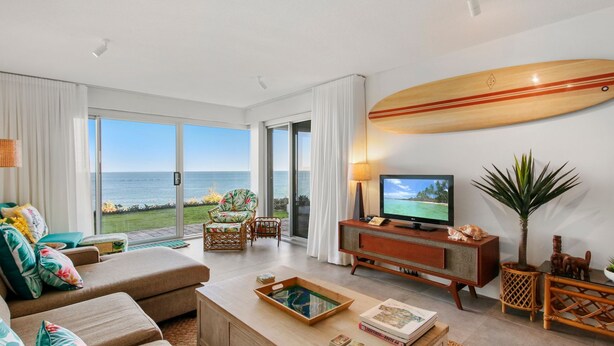 A spacious living room with a large TV, a surfboard decoration, and a view of the ocean. - Seaspray Unit 2, 21 Warne Tce, Kings Beach (Kings Beach)