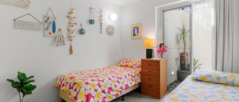 A bedroom with a bed, a dresser, a bedside table with a lamp, a wall-mounted light, and a window with curtains.