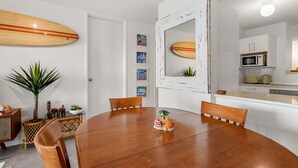 A dining area with a wooden table, chairs, and a surfboard decoration on the wall. - Seaspray Unit 2, 21 Warne Tce, Kings Beach (Kings Beach)