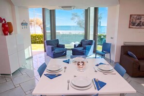 A dining area with a table set for four, two blue armchairs, a brown sofa, and a view of the sea through large windows.