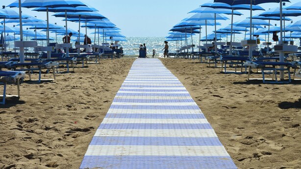 Een strand met een geribbelde mat naar het water, blauwe parasols en strandstoelen.