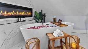 A modern bathroom with a large bathtub, a fireplace, and a wooden stool with towels.
