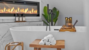 A modern bathroom with a freestanding bathtub, a fireplace, and a small wooden table with towels.