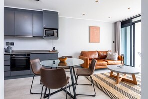 A modern kitchen with grey cabinets, a microwave, and a coffee machine. There is a dining area with a glass table and brown chairs. A brown leather sofa is in the living area with a view of the outdoors. - Stunning 204 Bellagio on Umhlanga Ridge (uMhlanga)